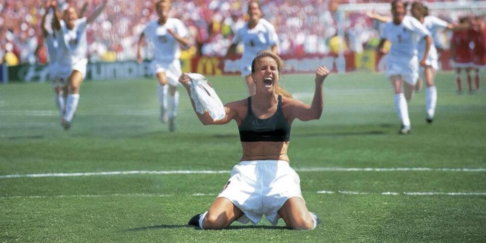 A woman is kneeling on a soccer field with her arms in the air.