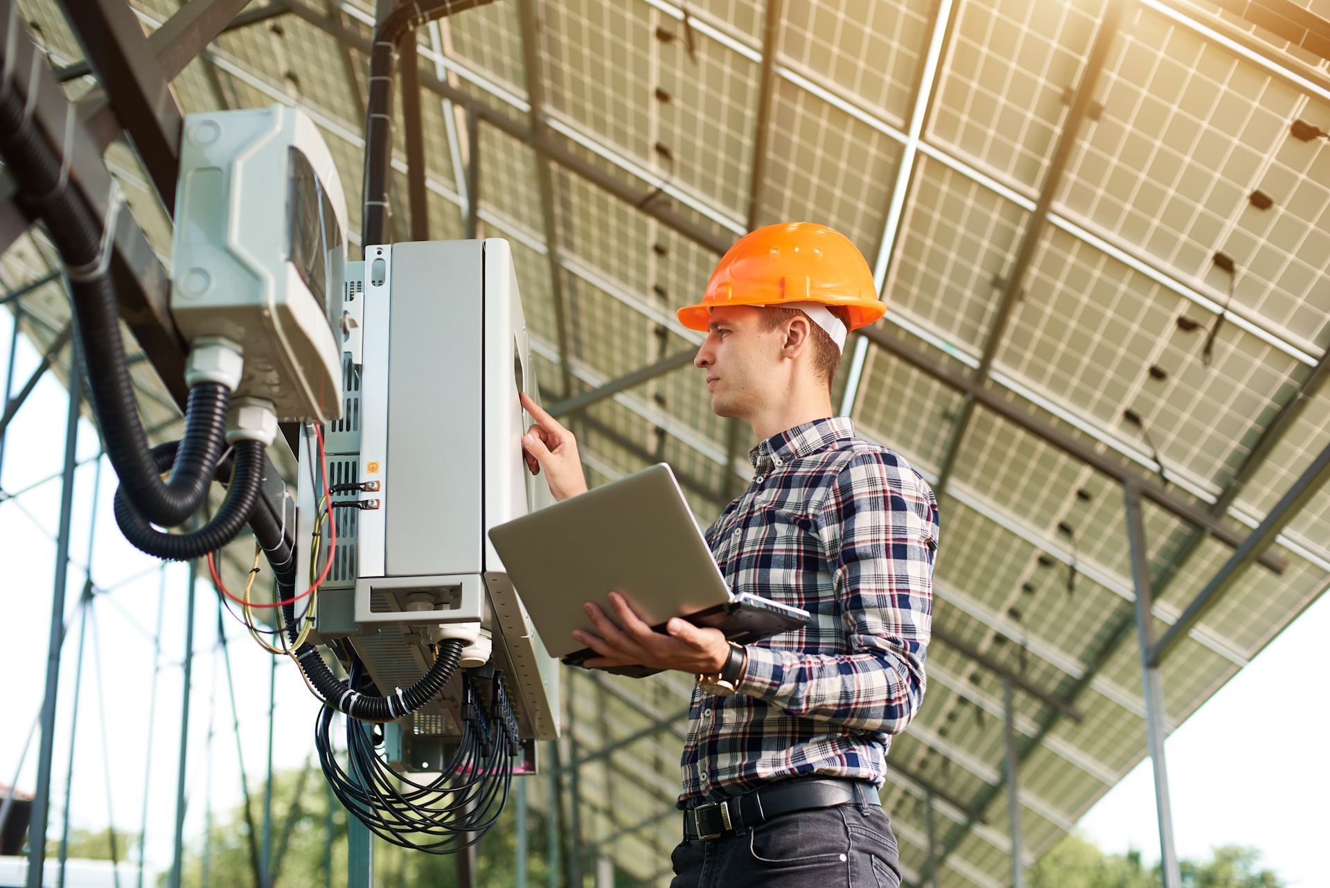 A man in a hard hat is working on a solar panel while holding a laptop.