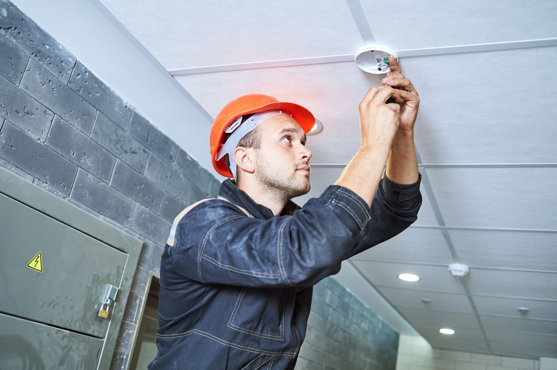 A man in a hard hat is installing a smoke detector on the ceiling.