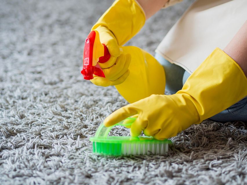 A person wearing yellow gloves is disinfecting a carpet with a spray bottle and a brush.