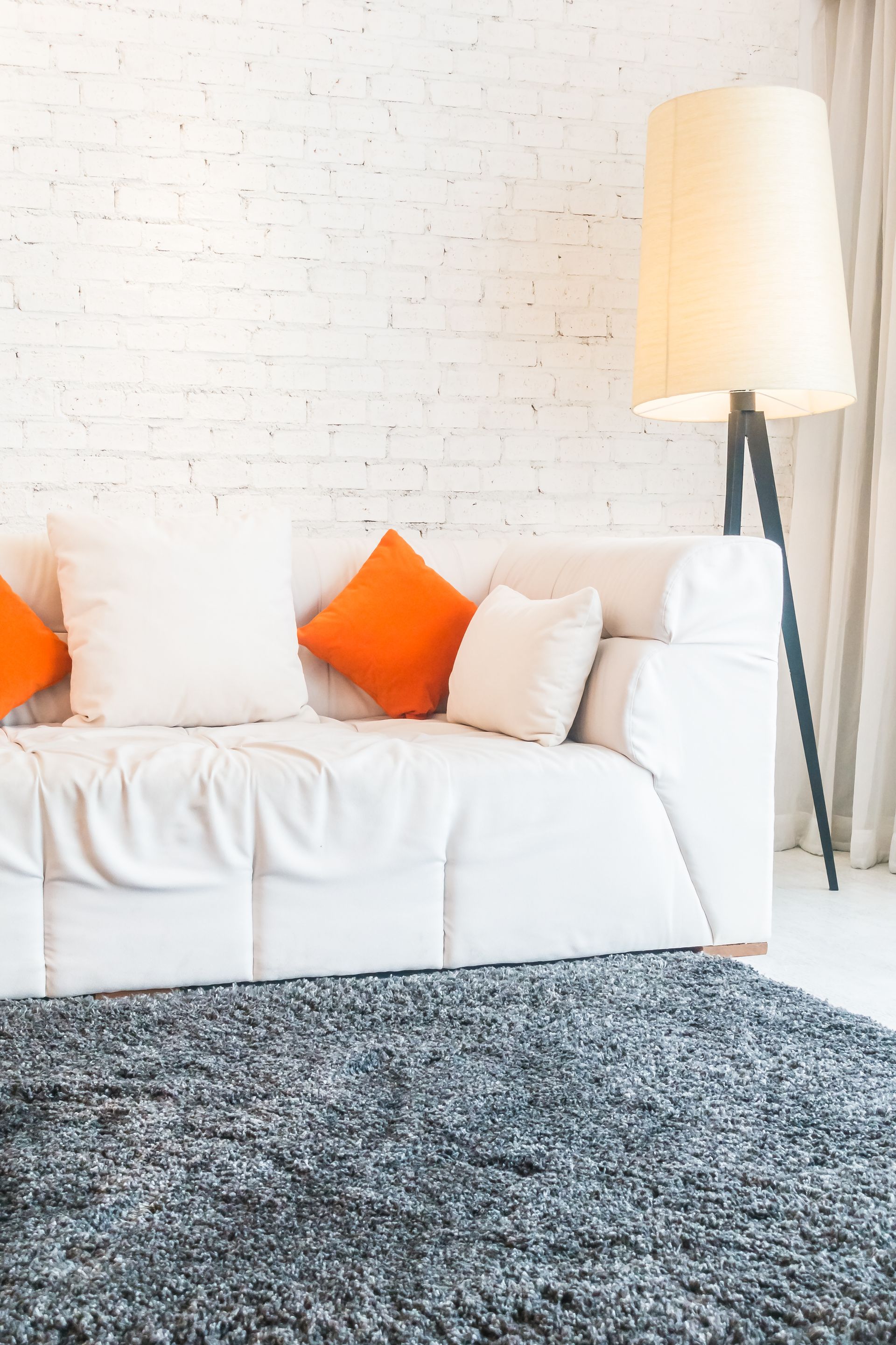 White couch with orange and white pillows, next to a tall lamp, set against a white brick wall and gray carpet.
