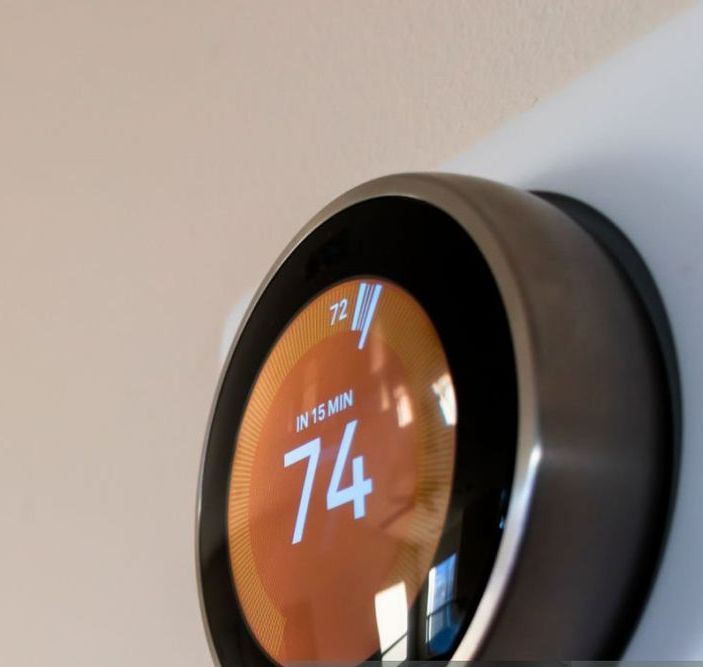 Close-up of a smart thermostat on a beige wall. The screen displays a temperature of 74 degrees.