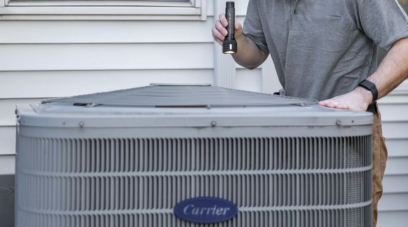 Person inspecting a Carrier air conditioning unit with a flashlight outside a house.