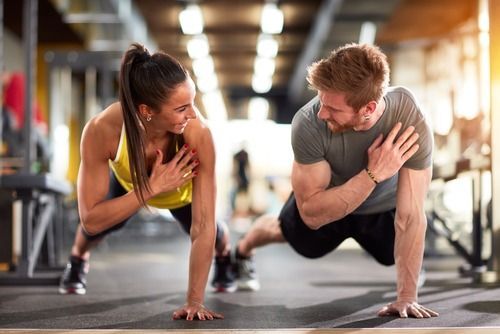 Woman and man doing push-ups in a gym, looking at each other with smiles, hands on chests.