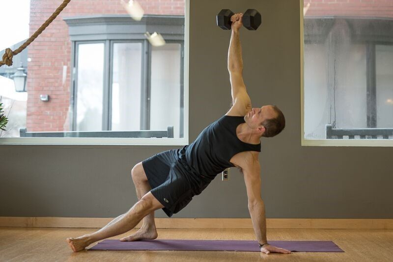 Man in black outfit doing a side plank with dumbbell overhead on purple mat.