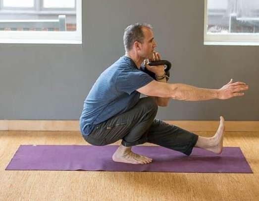 Man balancing in pistol squat, holding kettlebell, on yoga mat.
