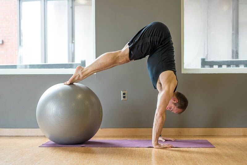 Man in handstand, feet on exercise ball, indoors on purple mat.