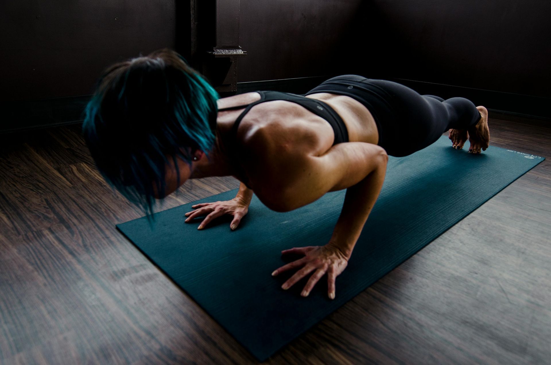 Woman with blue hair doing a push-up on a yoga mat. Dark room setting.