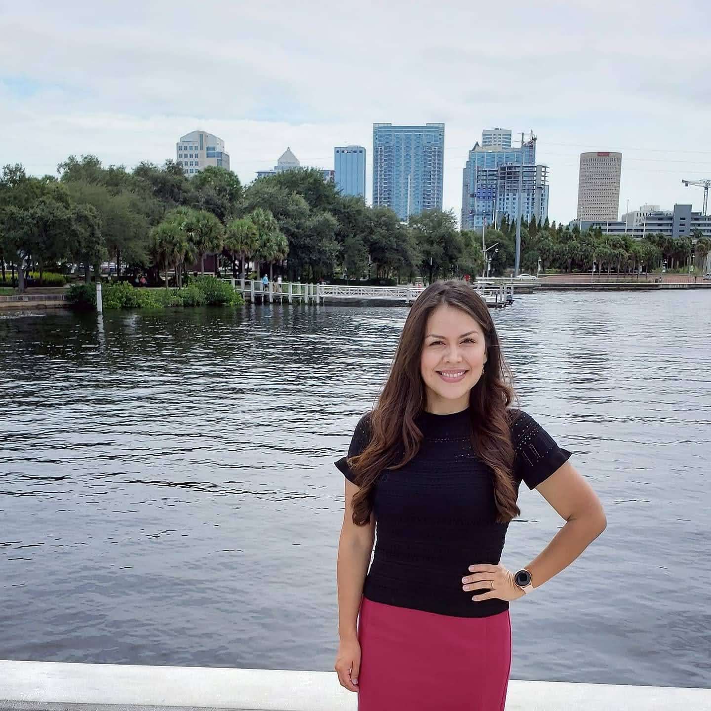 Realtor Esperanza in black top and pink skirt standing by a waterfront with city buildings in the background