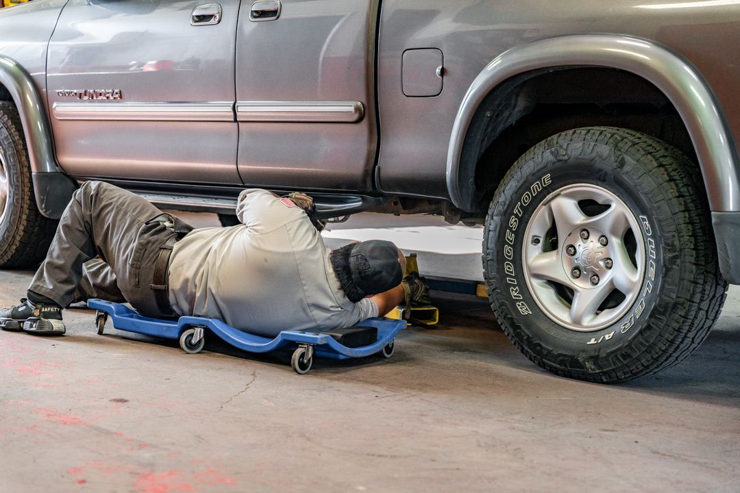 Mechanic lying on a creeper under a brown SUV, working near the rear wheel in a garage