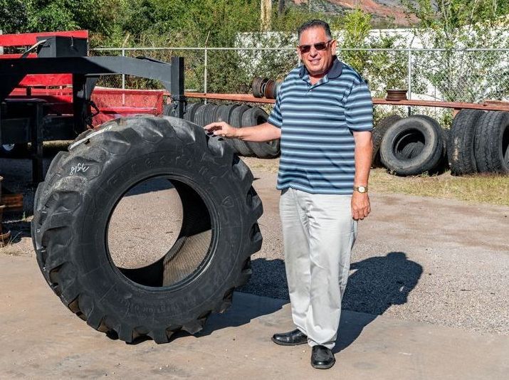 Man standing beside a giant black tire outdoors on a sunny day, with more tires and machinery behind him