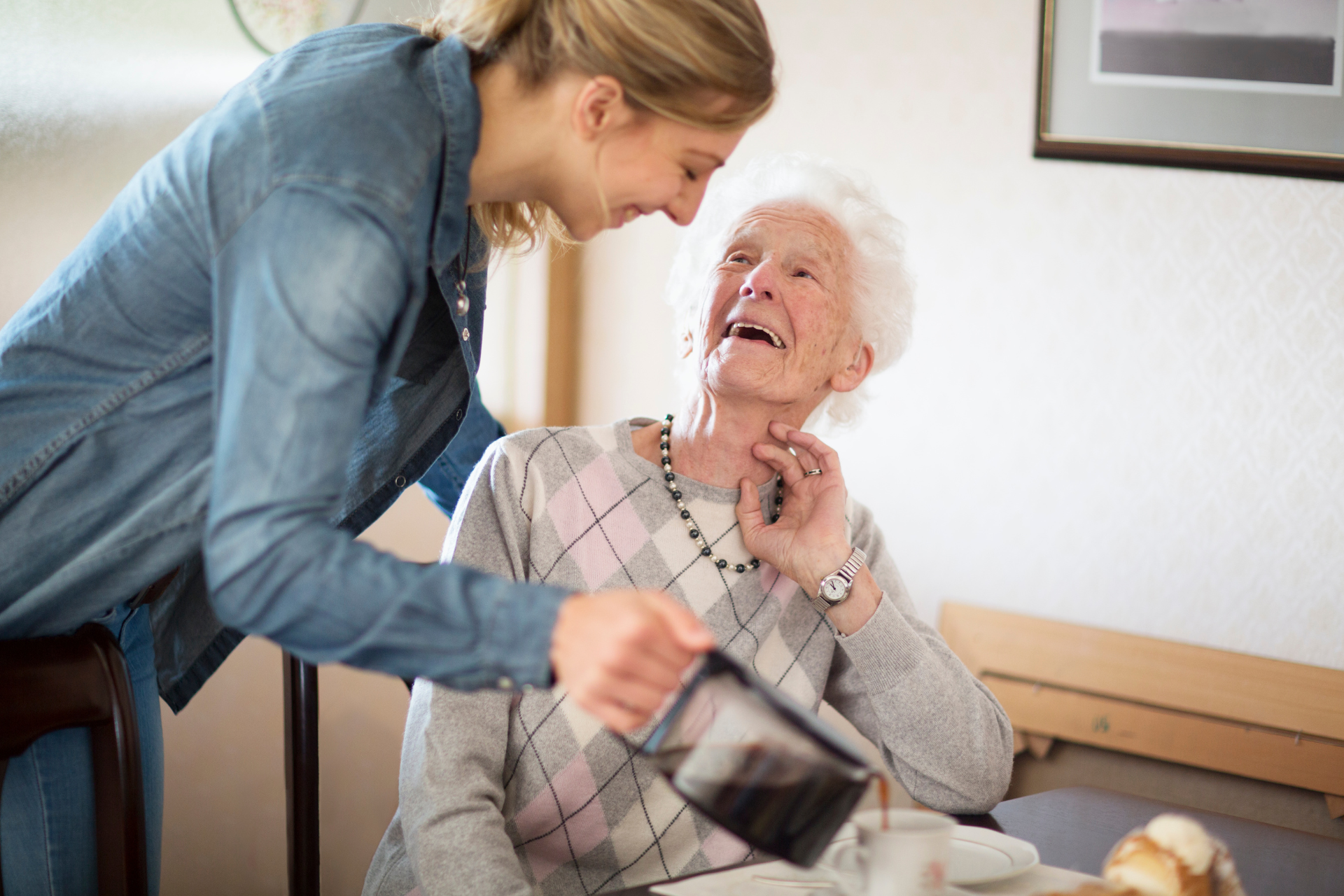 Caregiver helping an elderly woman stand beside a bed in a bright room