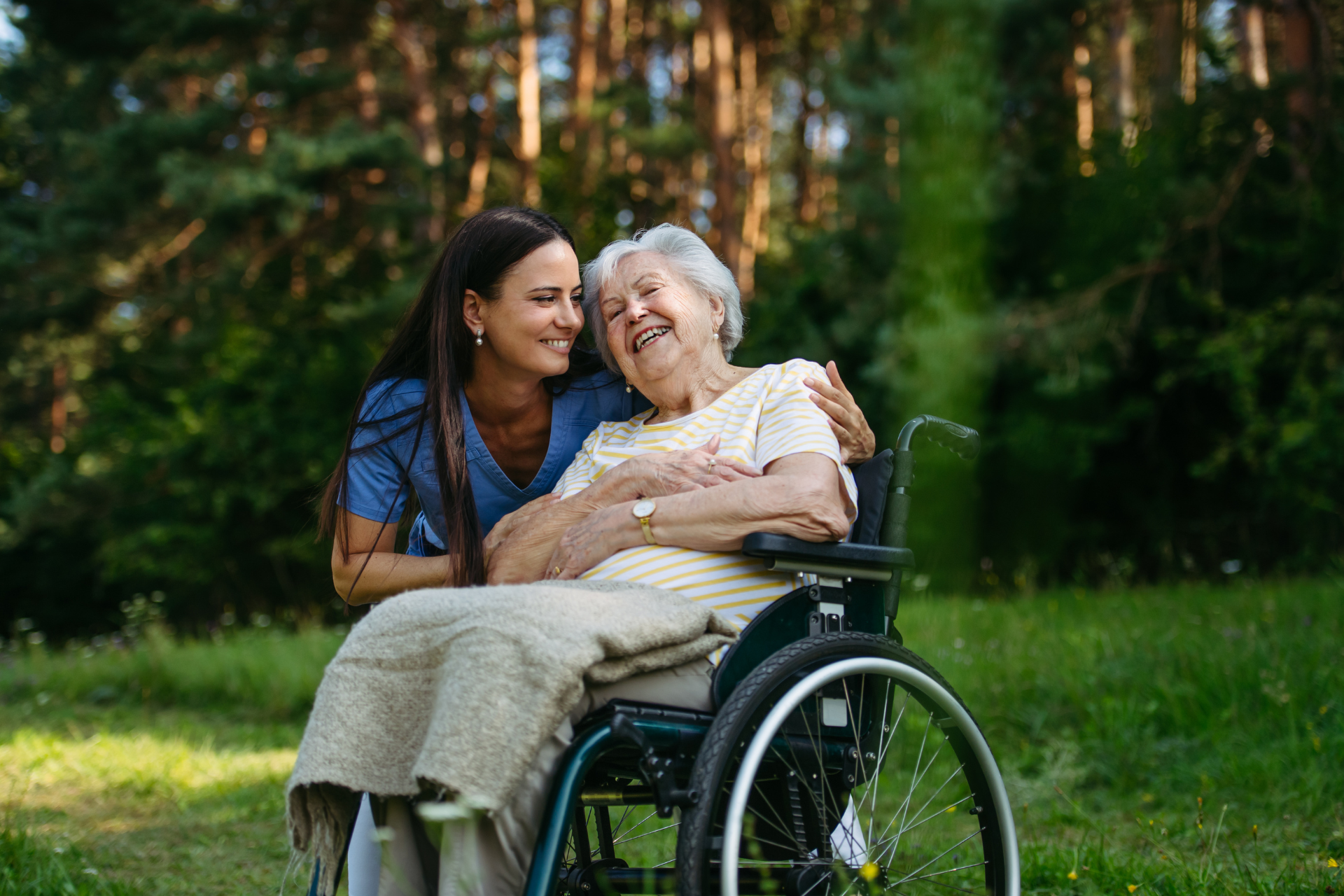 Young woman smiling beside an elderly person in a wheelchair outdoors on a grassy lawn