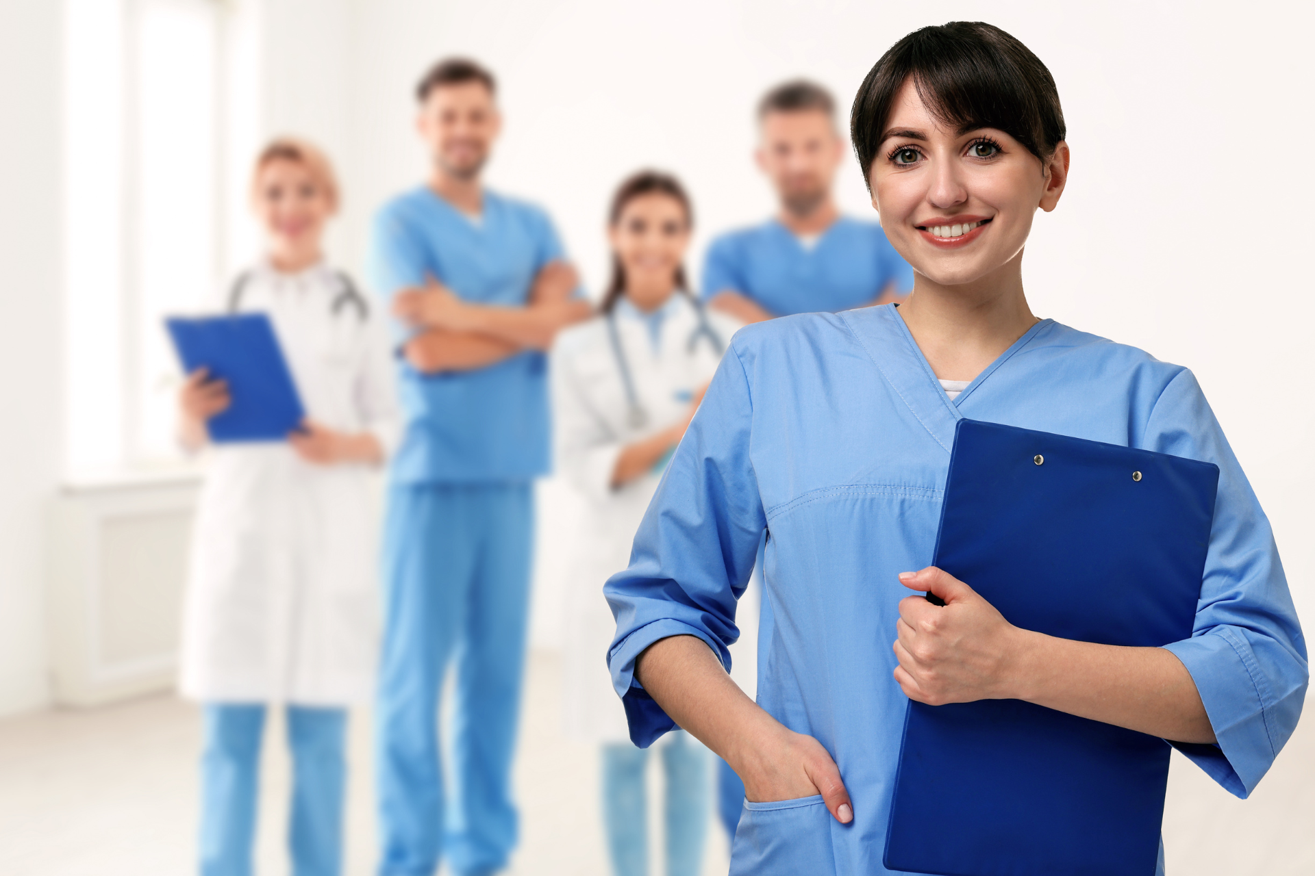 Healthcare team in blue scrubs standing in a bright hospital hallway, with a woman holding a clipboard in front.