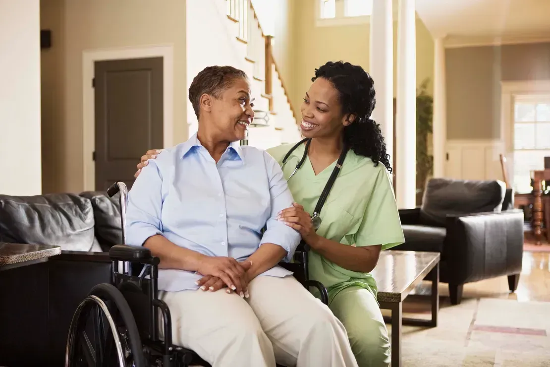 Caregiver smiling with seated patient in a living room beside a wheelchair