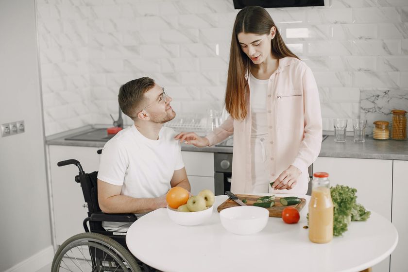 A woman is standing next to a man in a wheelchair in a kitchen.
