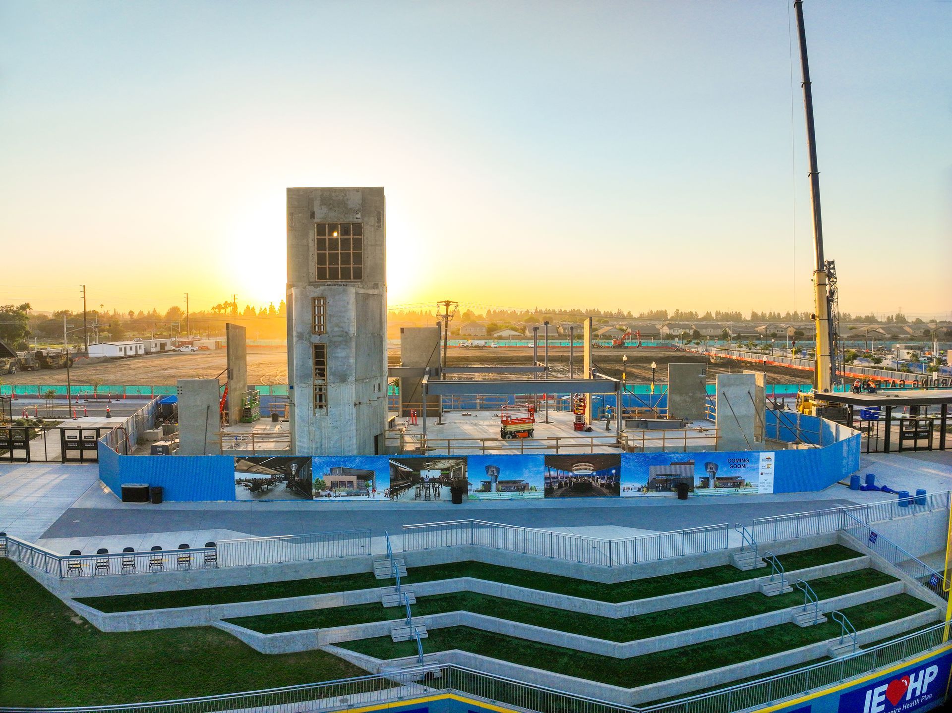 Sunset over a large industrial construction site with cranes, blue structures, and a tall central tower