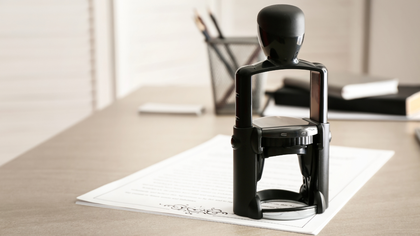 A black self-inking rubber stamp sits on a sheet of paper on a desk, with a pen holder and notebooks in the background.