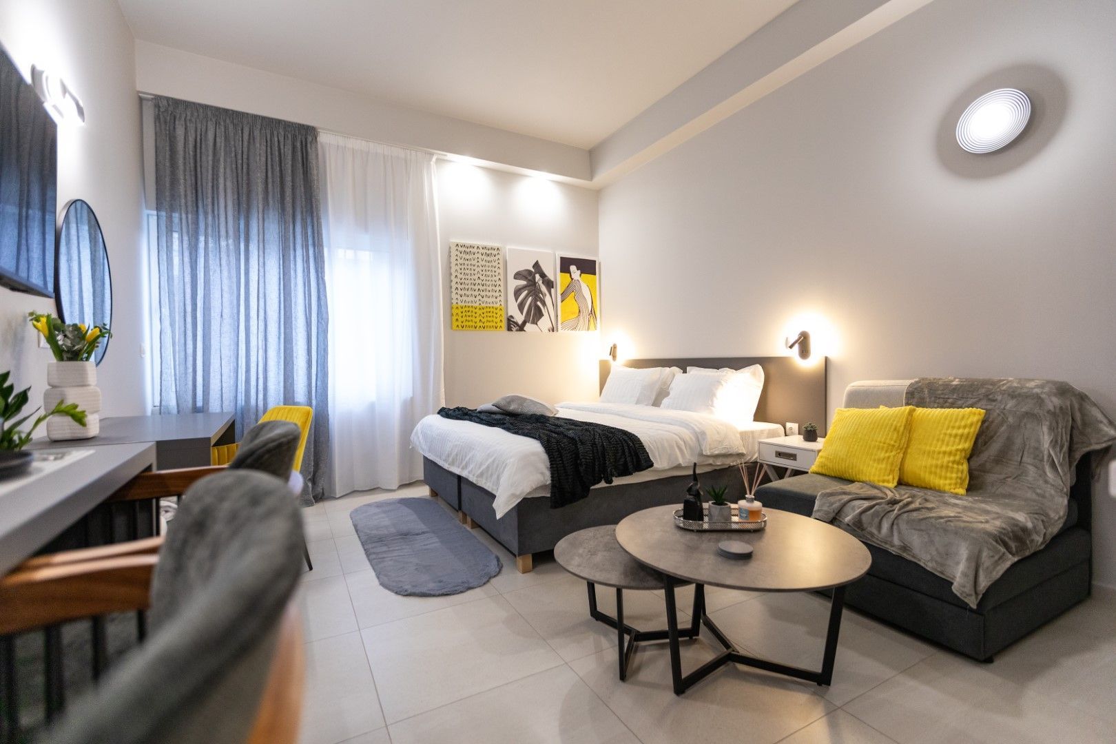 Modern hotel room with bed, sofa, desk, and round coffee table. Gray, white, and yellow accents.