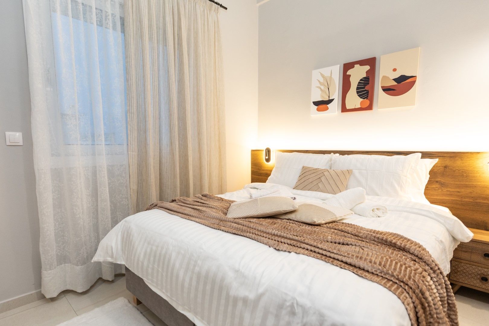 Bedroom with a bed, white bedding, brown throw, neutral wall art, and sheer curtains.