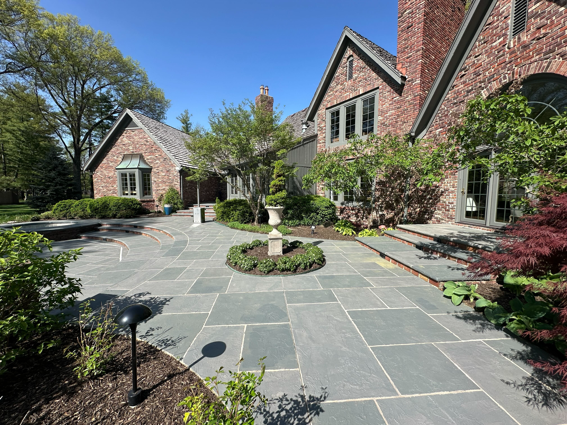 Brick home with a blue stone patio, a small fountain, and lush landscaping on a sunny day.