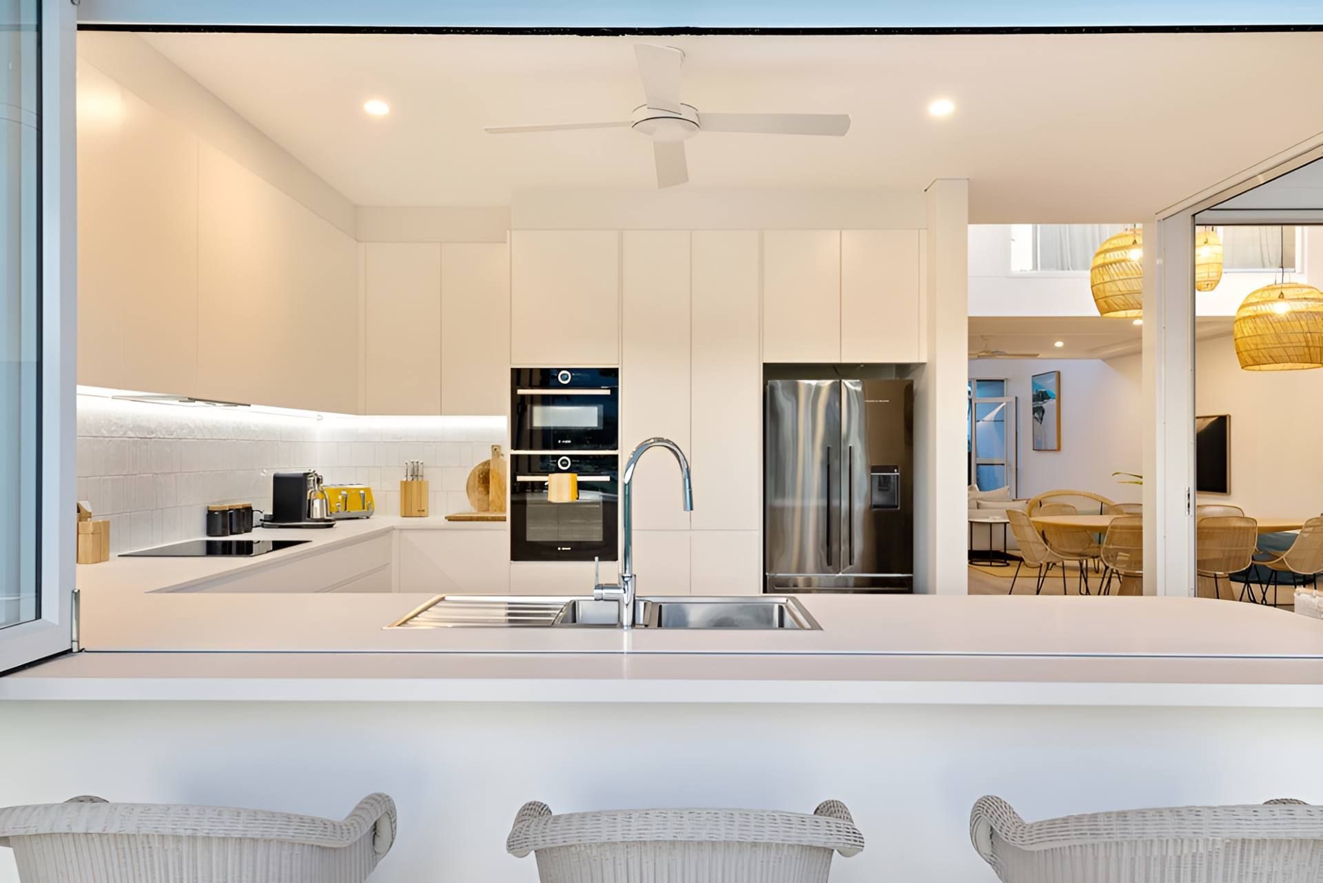 Modern White Kitchen With Island, Built-in Oven, Stainless Fridge, And Dining Area Visible Beyond — Kitchens by CCJS in Bungalow, QLD