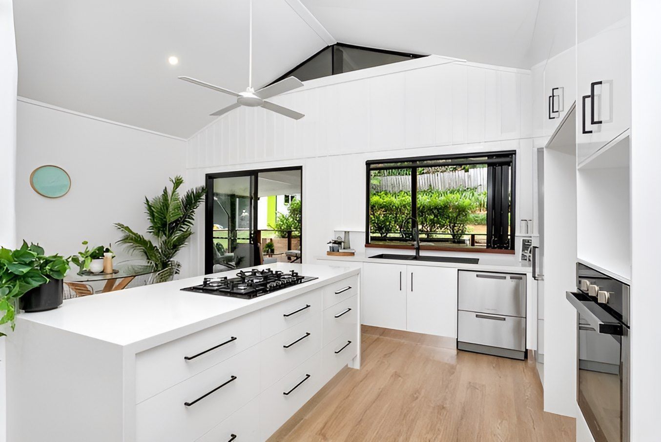 Bright Modern Kitchen With White Cabinets, Island, Wood Floors, And Large Windows Looking Outside — Kitchens by CCJS in Bungalow, QLD
