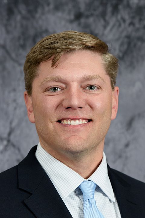 Studio headshot of a smiling person in a dark suit, light blue shirt, and tie against a gray background