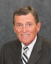 Studio portrait of a man in a dark suit, white shirt, and striped tie, smiling against a gray background