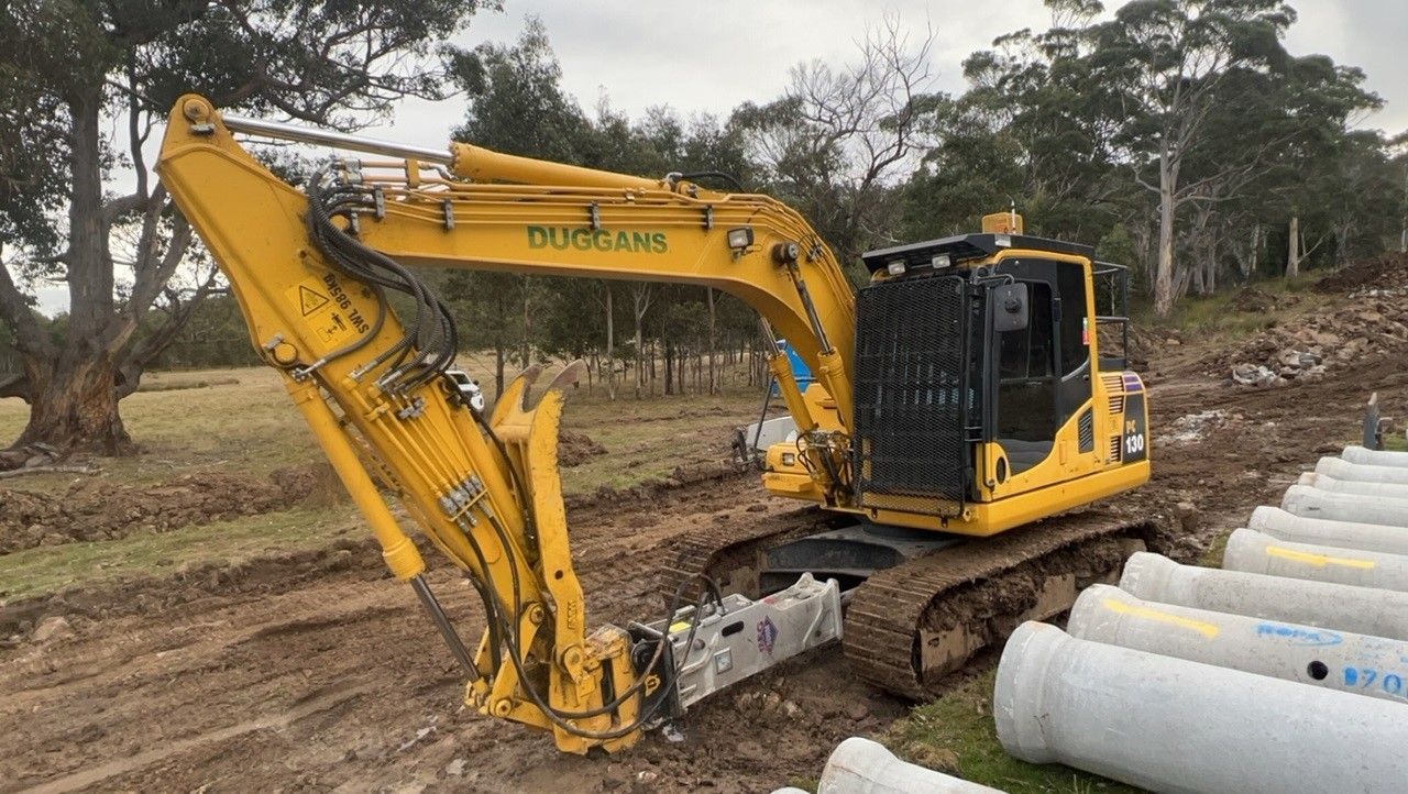 Yellow Komatsu excavator parked on a dirt road beside a blue truck and green bins under trees