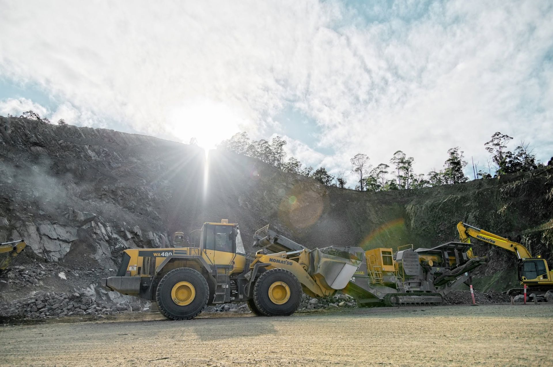 Yellow construction loader and conveyor machine on a gravel site with rocky hillside and bright sun glare