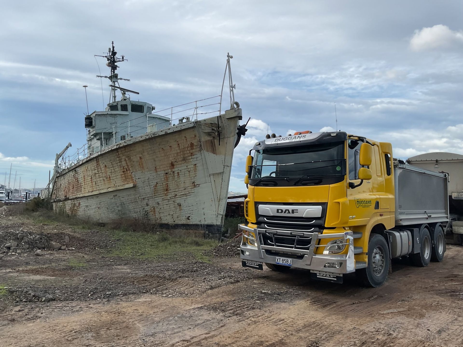 Yellow dump truck beside a rusted ship on a muddy shoreline under a cloudy sky