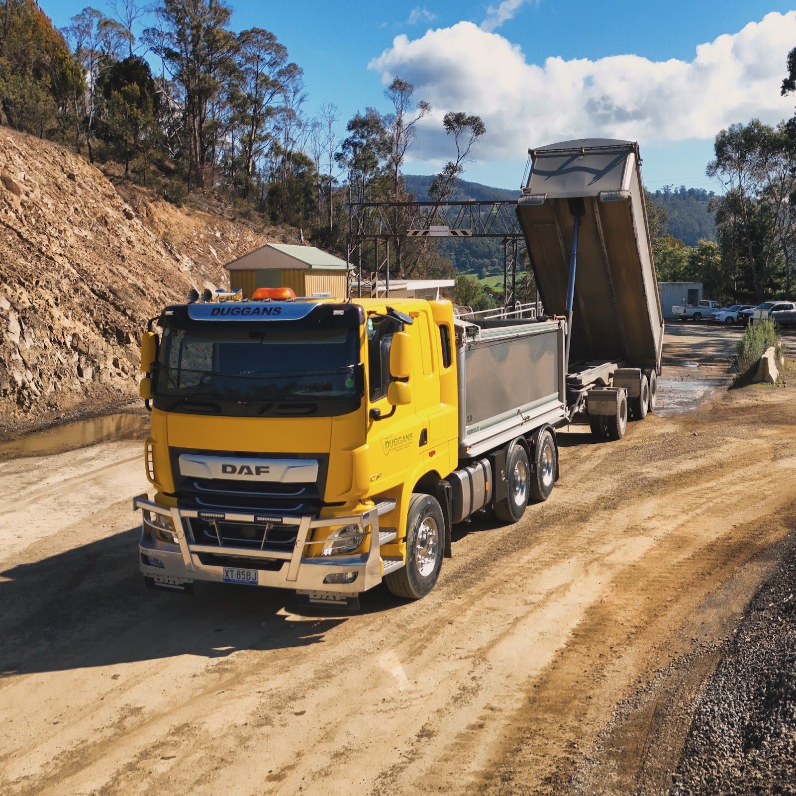 Crane lifting a long load on a flatbed truck beside a gravel road under a clear blue sky