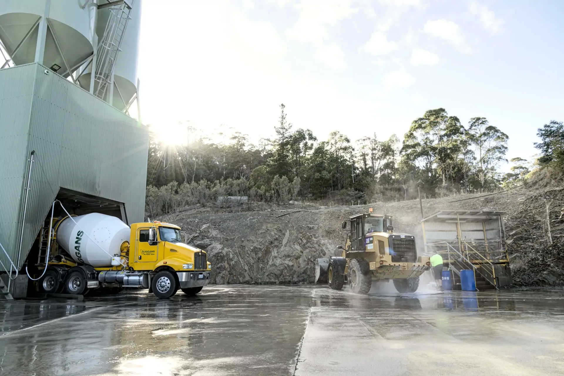 Yellow cement truck and loader at a wet construction site with a rock wall and water spray
