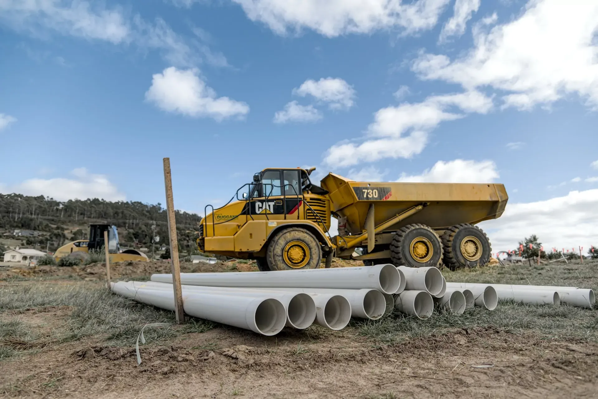 Yellow dump truck at a construction site beside stacked concrete pipes under a cloudy sky