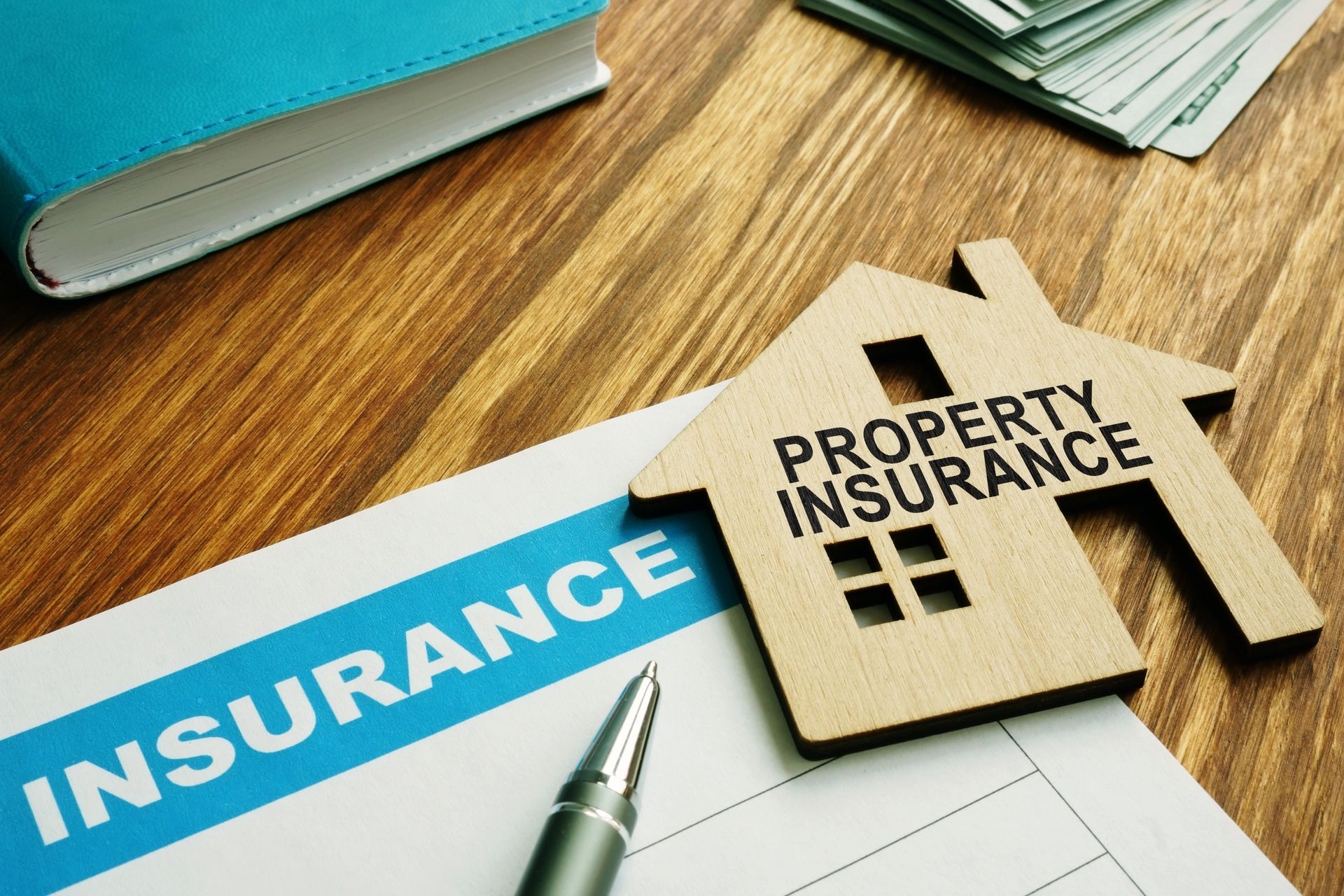 Property insurance paperwork with a house-shaped wooden cutout, pen, money, and a blue book on a wooden surface.