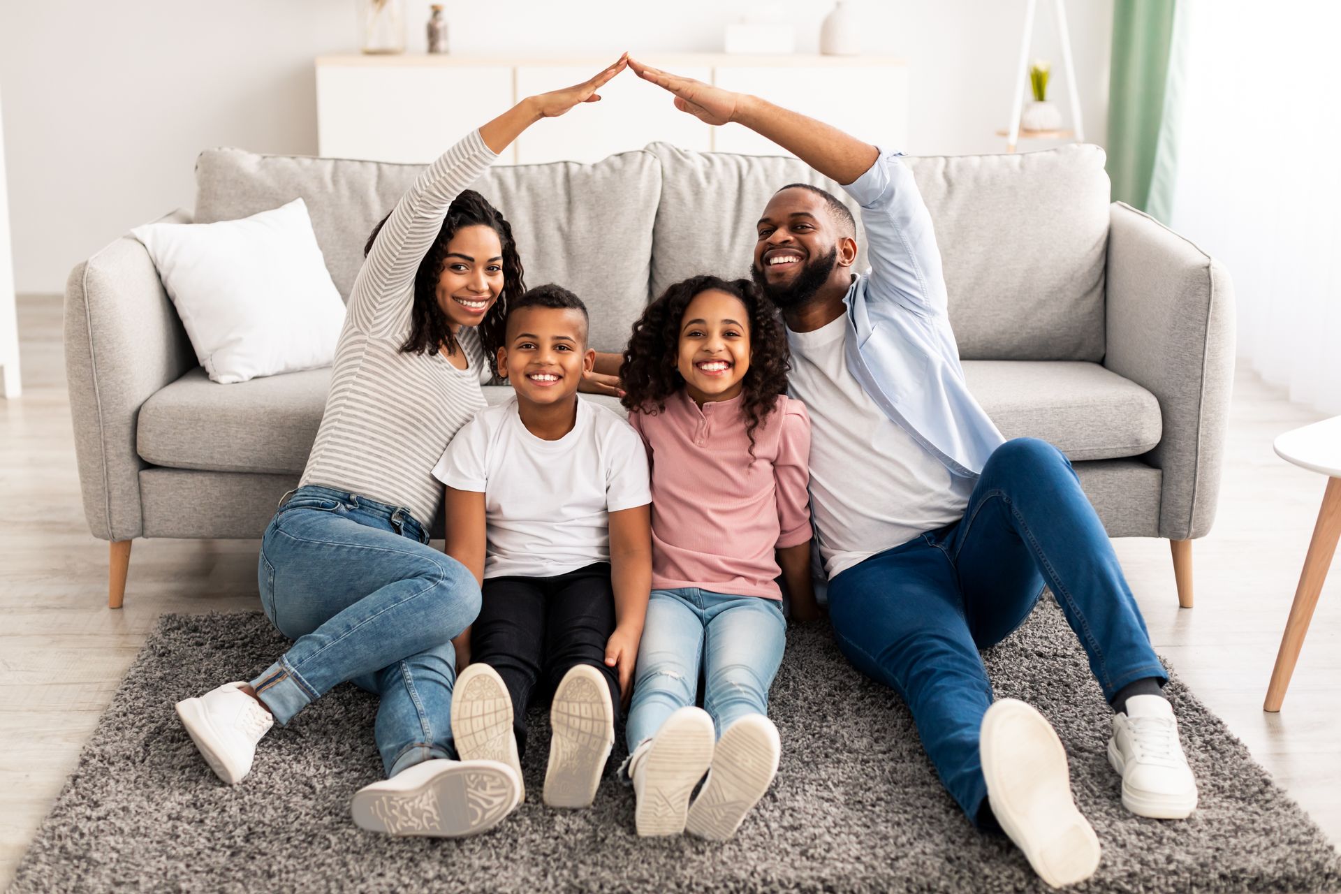 Family of four sitting on a rug in a living room, parents making a roof with their arms above their children's heads.