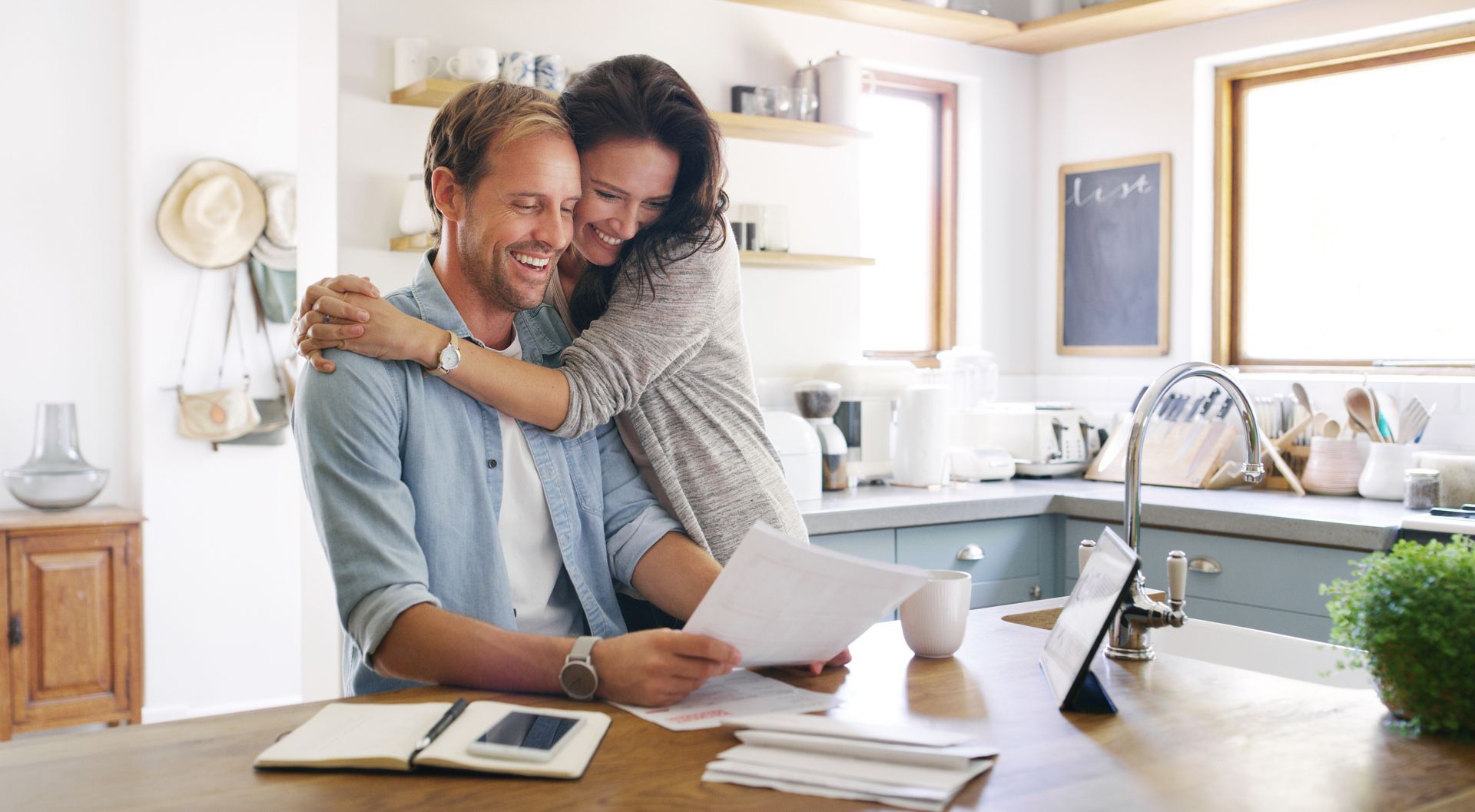 Couple smiling, embracing over papers and a tablet in a kitchen.