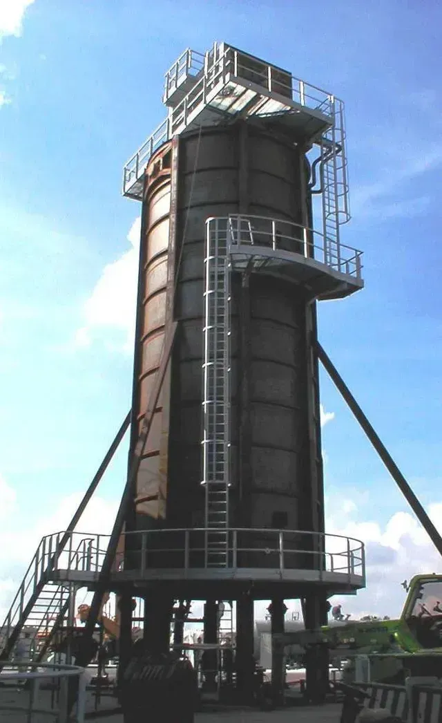 Un alto serbatoio industriale cilindrico con una scala e piattaforme, sostenuto da gambe, contro un cielo blu.