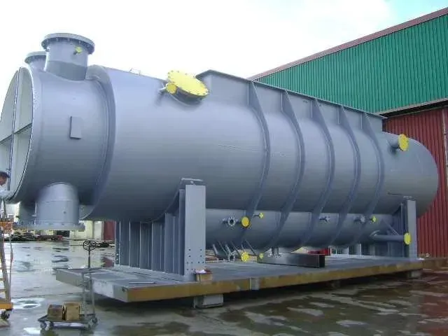 Grande nave industriale, grigia, su una piattaforma di trasporto. Tappi di scarico gialli. Ambientazione esterna.