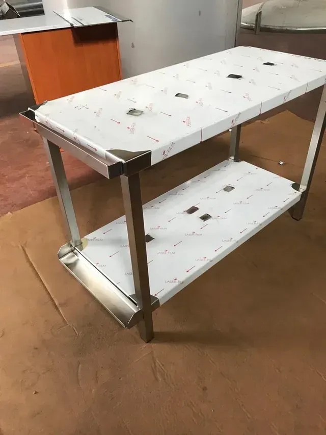 Tavolo da lavoro in acciaio inossidabile con ripiano inferiore, appoggiato su una superficie marrone.