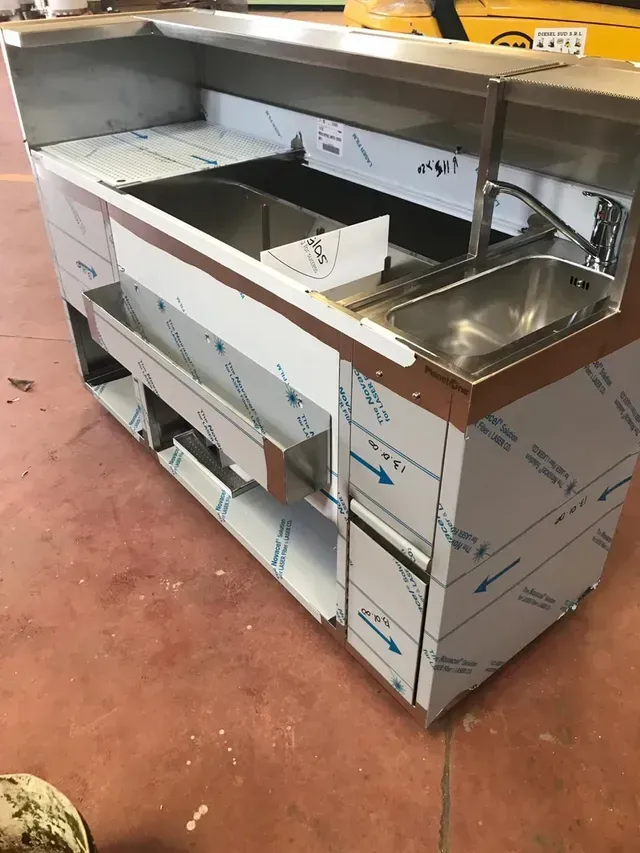 Bar commerciale in acciaio inox con lavello, vaschette per il ghiaccio e spazio di archiviazione, su pavimento rosso.