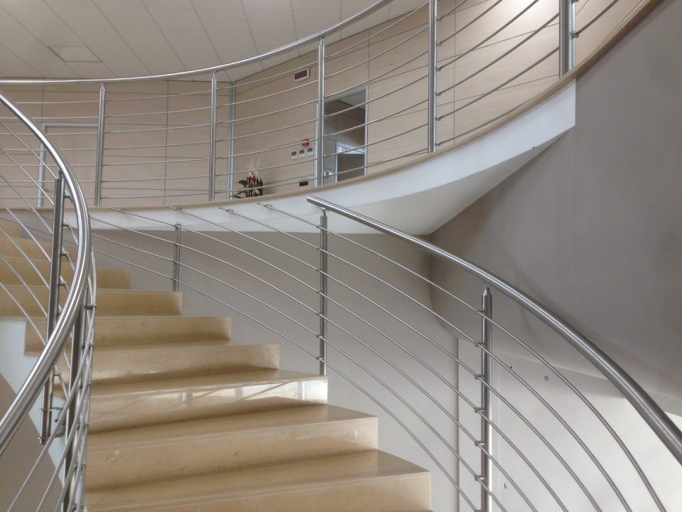 Scala curva con corrimano in acciaio inox e gradini in pietra chiara, che sale verso un livello superiore.