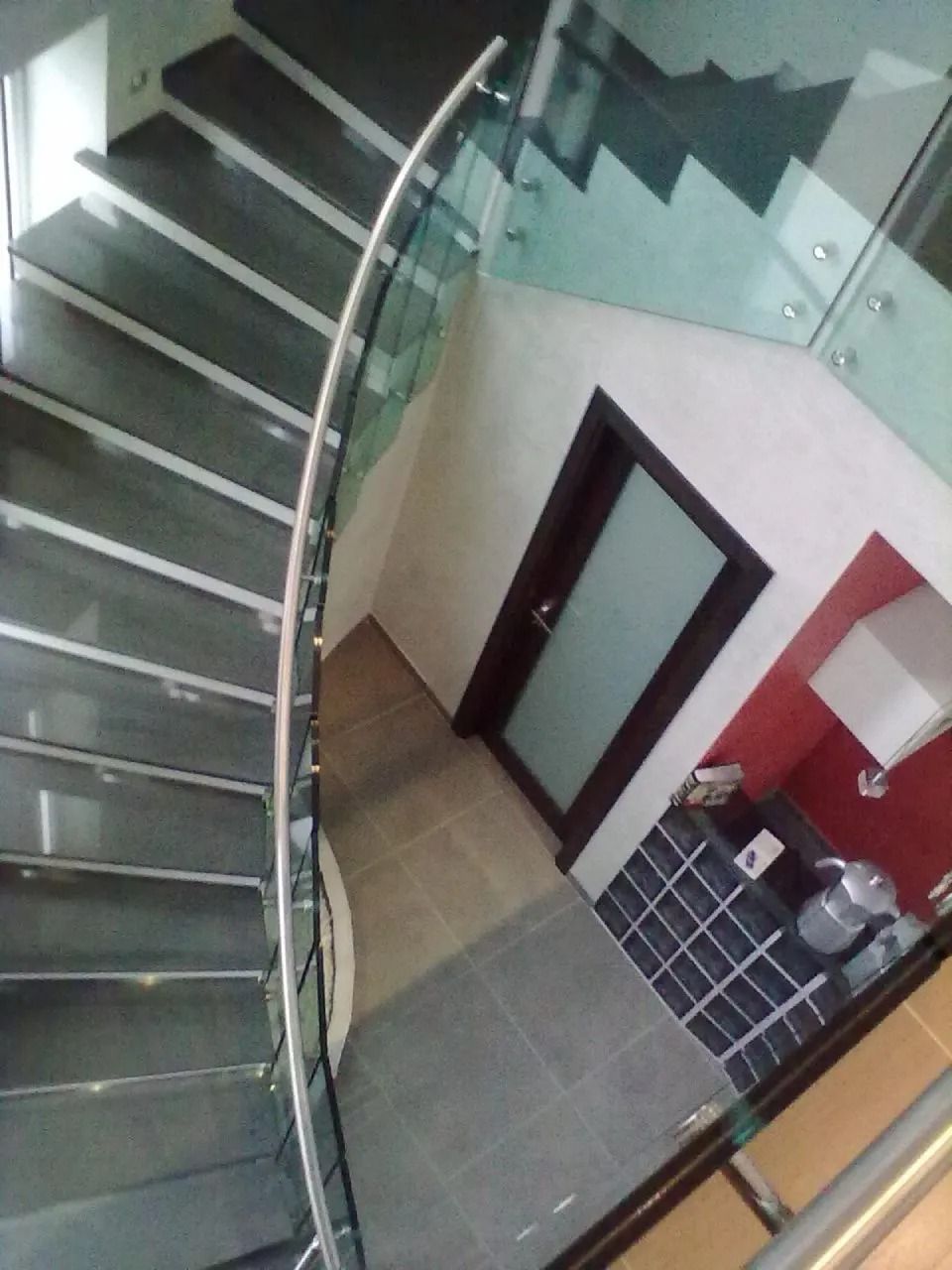 Scala curva con ringhiera in vetro. Gradini e pavimenti grigio scuro conducono a una porta e a una parete scura.