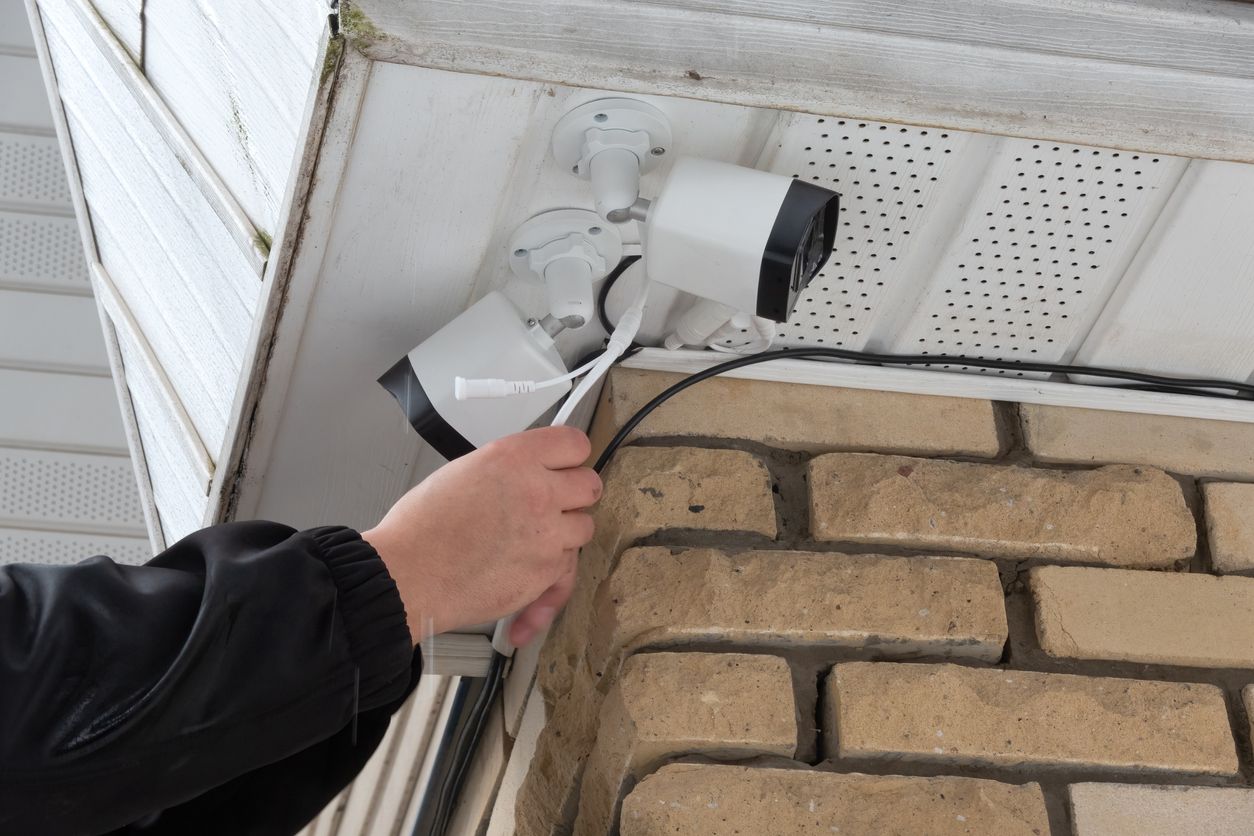 Hand holding a silver security camera mounted under white siding beside a brick wall