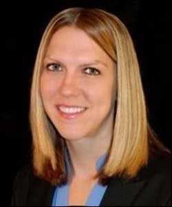 Portrait of a smiling blonde woman in a black blazer and blue top against a dark background