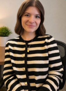 Smiling person in a black-and-cream striped sweater seated indoors beside a small plant.