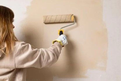Person painting a beige wall with a paint roller, covering a patchy area.