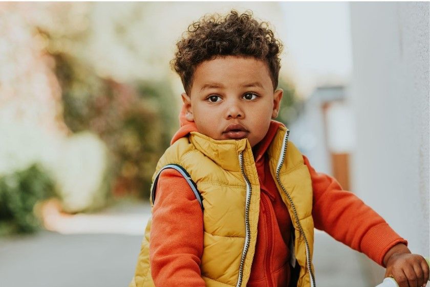Young child in a yellow vest and orange shirt standing outdoors, looking at the camera.