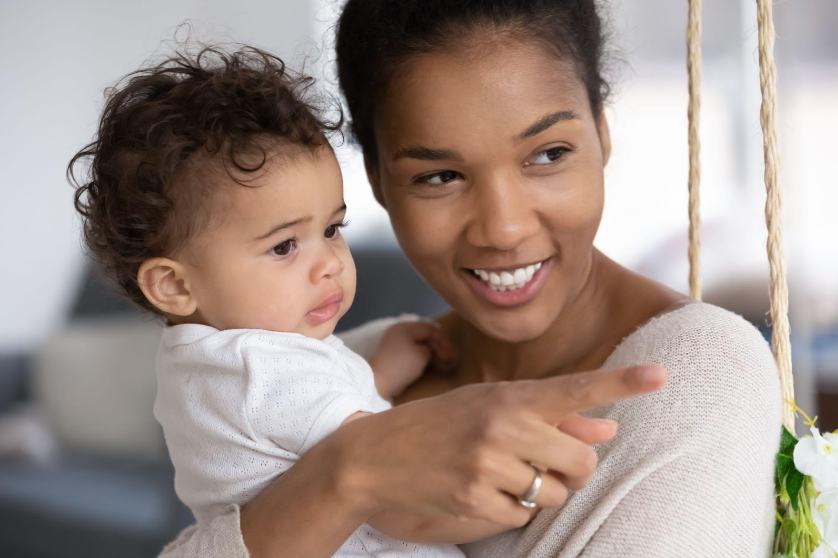 Smiling parent holding a baby and pointing at something offscreen in a bright room